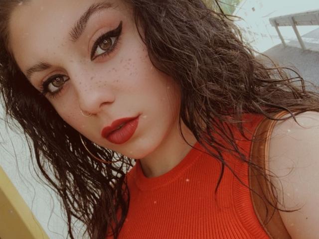 Butterflysophia69 with wet, curly black hair, red lipstick, black eyeliner, freckles, nose ring, and red ribbed tank top, looking at camera with serious expression.