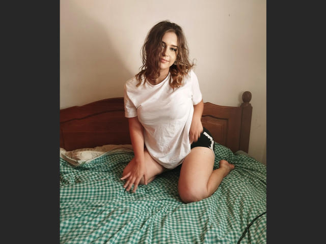 Sisilia, with wavy brown hair, wearing a white tee and black panties, sits on a green checkered bed, smiling, knees on bed, hands on bed, looking cute.