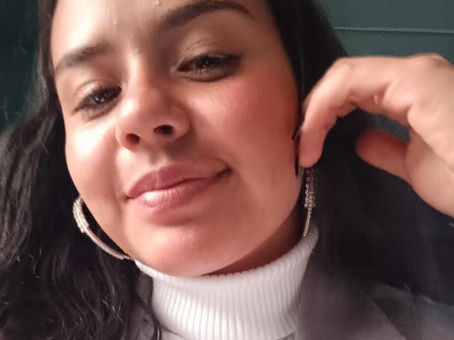 ArielleWilde's face with dark, wavy hair, brown eyes, and light skin, wearing a white turtleneck and silver hoop earrings, smiling slightly with a hand near her ear.