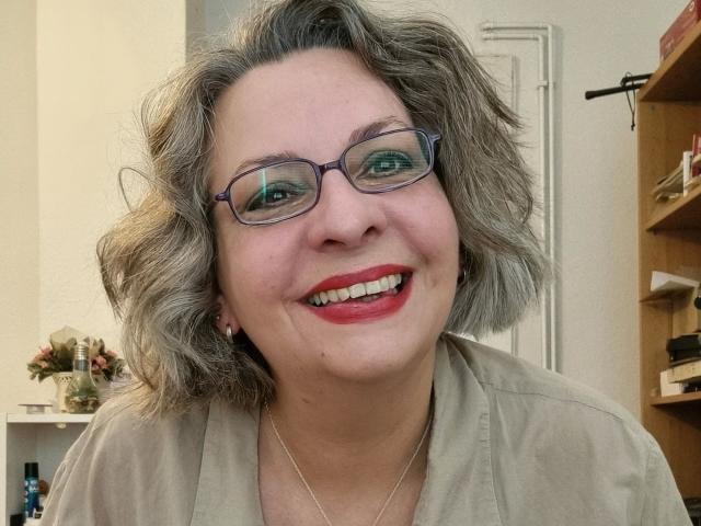 CashLadyVivian: una donna attraente con capelli grigi corti e ondulati, occhiali, rossetto rosso e un sorriso, indossa una camicia beige e una sottile collana, in un selfie..