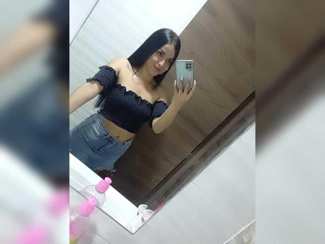 HikariChloe mit langen, schwarzen Haaren, trägt ein schwarzes, schulterfreies Crop-Top und blaue Jeans und macht ein Selfie vor einem Spiegel, lächelnd, wobei ihre kleinen Brüste und ihr hübsches Hinterteil zu sehen sind..