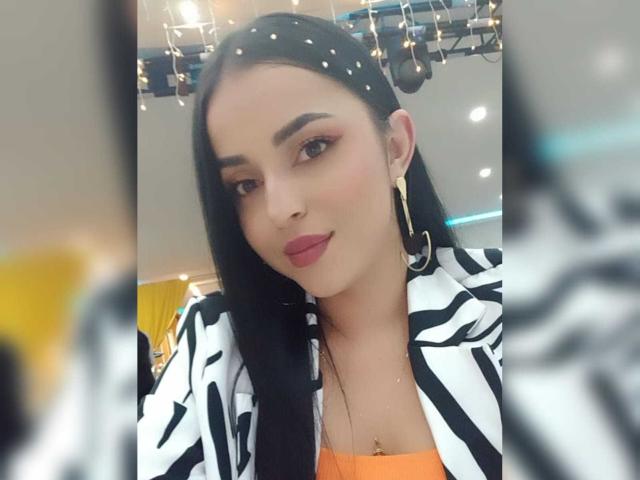 HikariChloe with long black hair, wearing a zebra-striped jacket, orange top, red lipstick, gold hoop earrings, and a small necklace, looking at the camera with a slight smile.