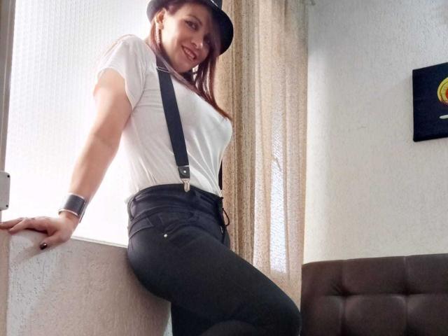 MilenaHott69 with brown hair, white t-shirt, black suspenders, black pants, smiling, wearing a black cap, black wristband, leaning against a wall, showing a cute butt.