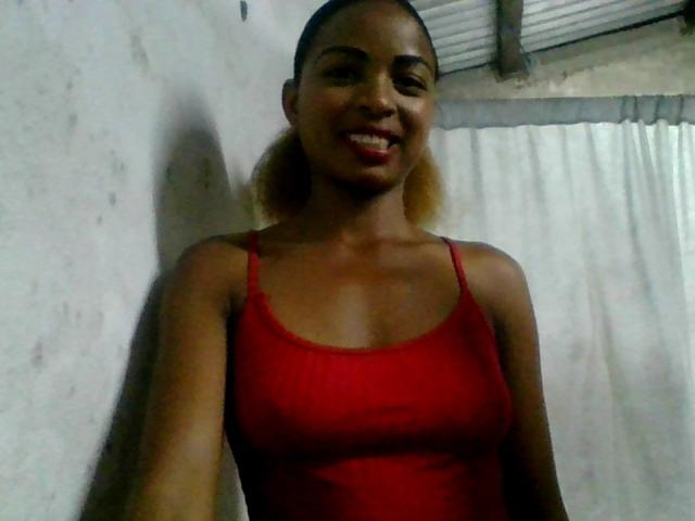 AlexaChan with dark skin, red tank top, small tits, blonde ponytail, smiling, standing against a wall, shadow on the left, under a corrugated roof with a white curtain.