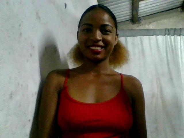 AlexaChan with dark brown skin, red tank top, and blonde puffy hair, smiling at the camera, showing her tits and ass in the photo.