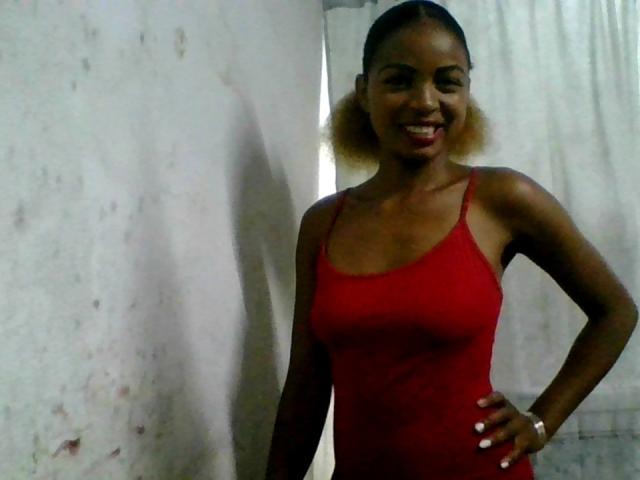 AlexaChan with dark skin, small tits, and a red tank top, smiling, hand on hip, light brown puffy hair, white nails, standing against a slightly dirty wall.
