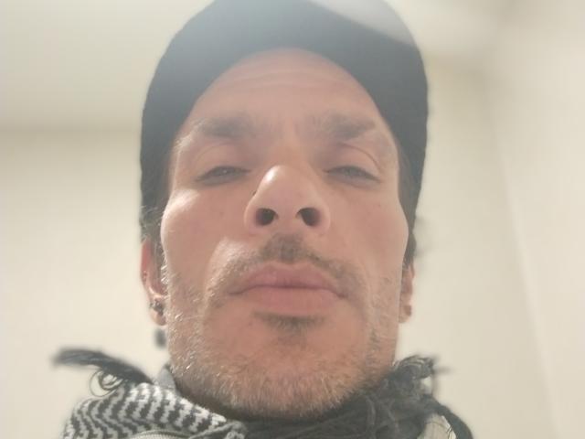 Chiroco has a serious face, a black beanie, and a gray beard with slight stubble. He's wearing a black and white striped scarf, looking straight at the camera.