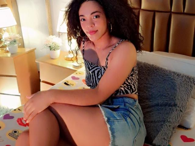EmilylLight with curly hair, zebra-striped top, showing her tits, in blue denim shorts, sitting on a bed with a gray pillow, smiling at the camera.