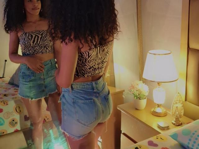 EmilylLight with curly hair, zebra print strapless top, and frayed denim skirt, standing in front of mirror, showing her ass and reflecting her face. Bedroom with lamp, flowers.