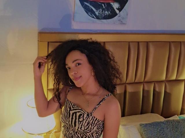 EmilylLight with curly black hair, wearing a zebra-patterned dress, smiling, holding her hair, standing by a bed with a gold headboard, and a lit lamp on the left.