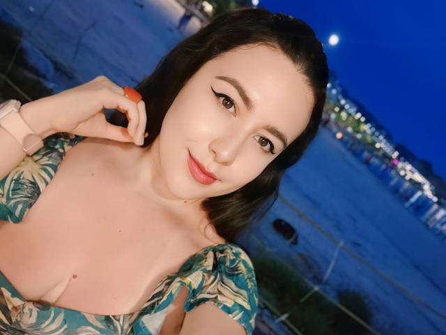 MabyLegrand with pale skin, dark hair, and red lipstick, holding an orange, wearing a green leaf-patterned dress, showing her tits, and a pink watch, under a blue night sky.