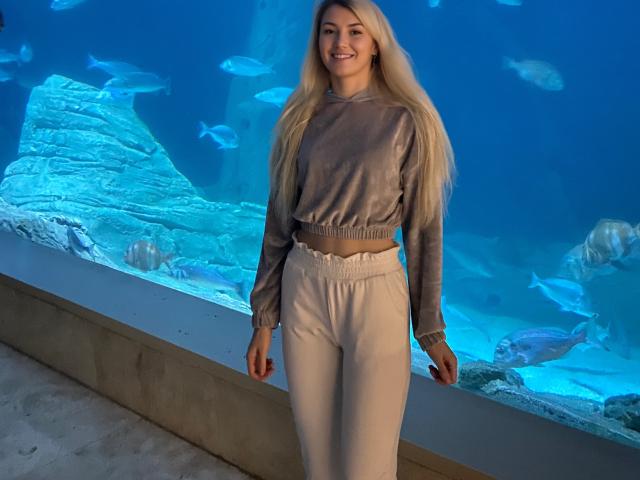 MoonBabeX with long blonde hair, smiling, wearing a gray sweatshirt and high-waisted white pants, standing in front of a blue aquarium with fish and rocks.