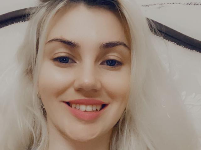 MoonBabeX has blonde hair, blue eyes, and a cute smile with pink lips. She's looking at the camera, showing her teeth. Her eyebrows are dark and she's looking happy.