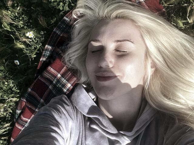 MoonBabeX, blonde woman with fair skin, lying on grass, eyes closed, soft smile, gray hoodie, red plaid blanket, sunlit, gentle shadows, wavy hair, relaxed, natural light.