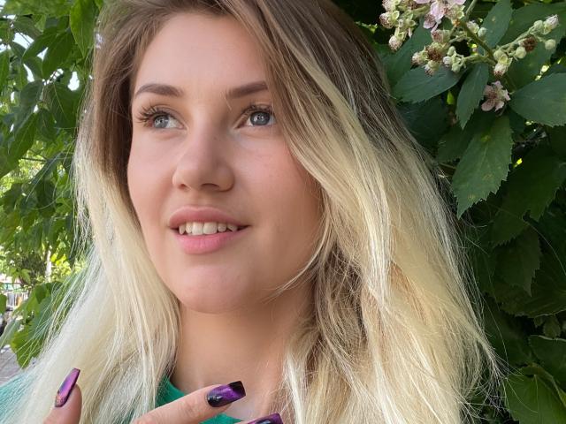 MoonBabeX has blonde, wavy hair, blue eyes, and purple nails, smiling with slightly parted lips, showing a cute, relaxed face. She's standing in front of green leaves.