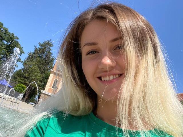 MoonBabeX has long blonde hair, a green shirt, and a cute smile, standing outside with a clear blue sky. Her ass is not visible.