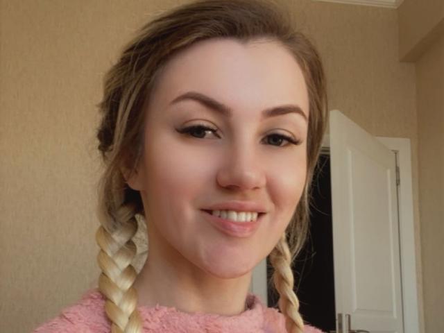 MoonBabeX has blonde braids, thick eyebrows, and a pink fluffy shirt, smiling with a slight nose chill, brown eyes, and a cute face.
