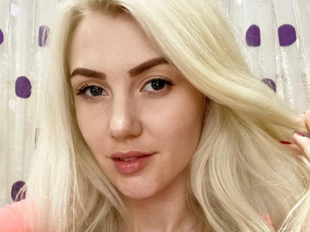 MoonBabeX has blonde hair, blue eyes, and a slight smile with pink lips. She's holding her hair with red-manicured fingers. Her eyebrows are thick and her skin is fair.