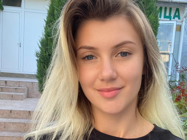 MoonBabeX with blonde, ombre hair, blue eyes, pink lips, and fair skin, smiling softly. She's wearing a black top, and her tits are small and cute.