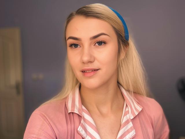 MoonBabeX has blonde hair, blue headband, pink shirt, striped white and pink shirt, and is smiling with light makeup and blue eyes. She's looking straight at the camera.
