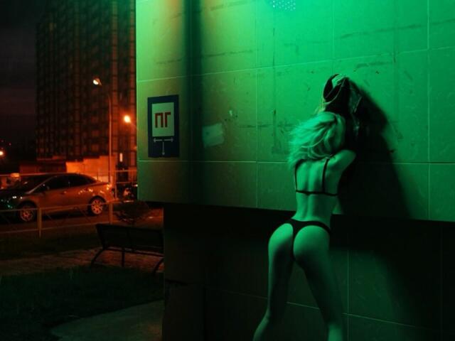 ElzaKomx with blonde hair, wearing black thong and bra, back to camera, pressing against green-lit tiled wall, under neon light, at night, with cars and building in background.