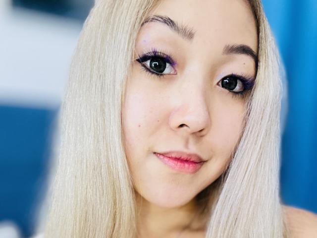 RenyLime a les cheveux blonds, du fard à paupières violet et de longs cils noirs. Elle sourit avec des lèvres roses et a une peau claire. Son visage est centré et elle regarde l'appareil photo..