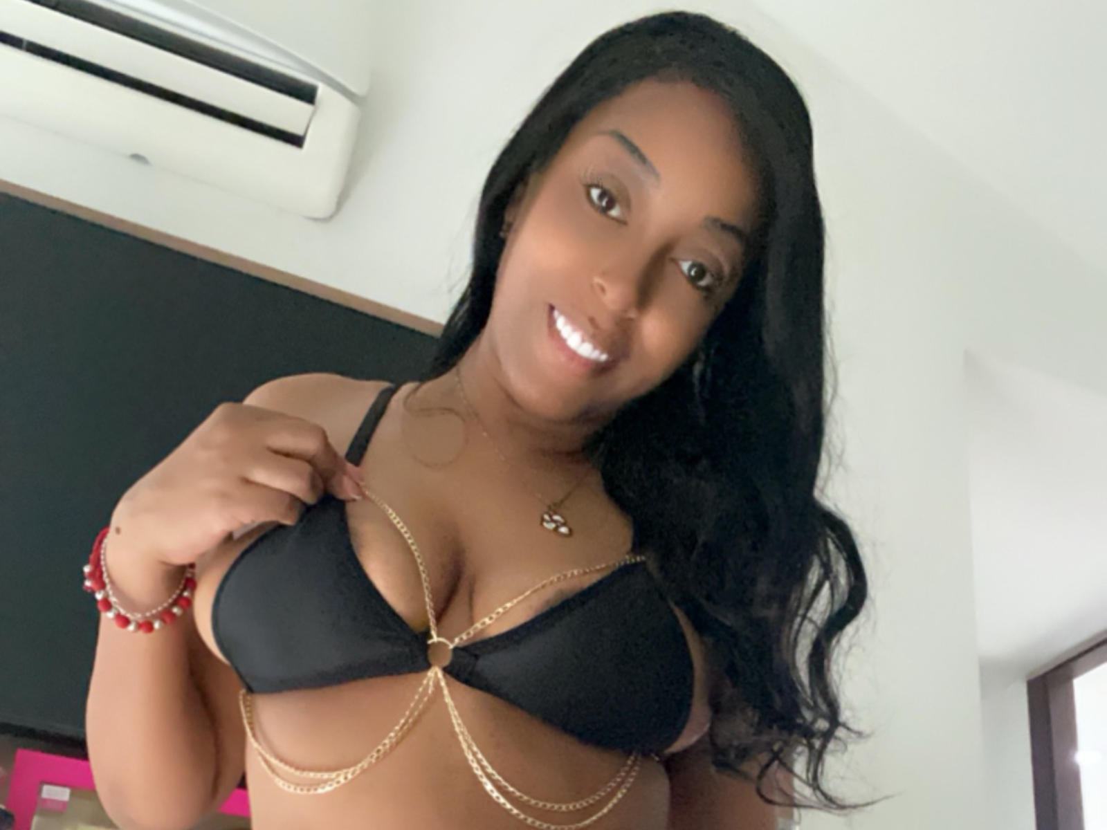 MatureBlack with dark skin, long black hair, smiling, wearing a black bra with gold chains, showing her boobs, red bracelet, and gold necklace.