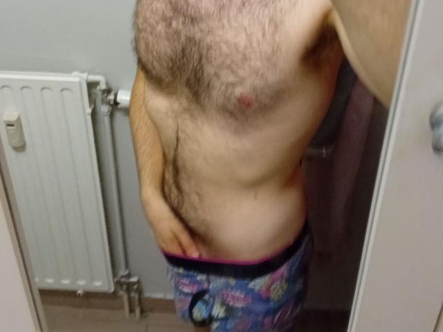 Il petto e l'addome pelosi di Aure69, con un piccolo capezzolo eretto, indossa pantaloni colorati con motivi floreali, è in piedi in una stanza e mostra una mano sull'inguine..