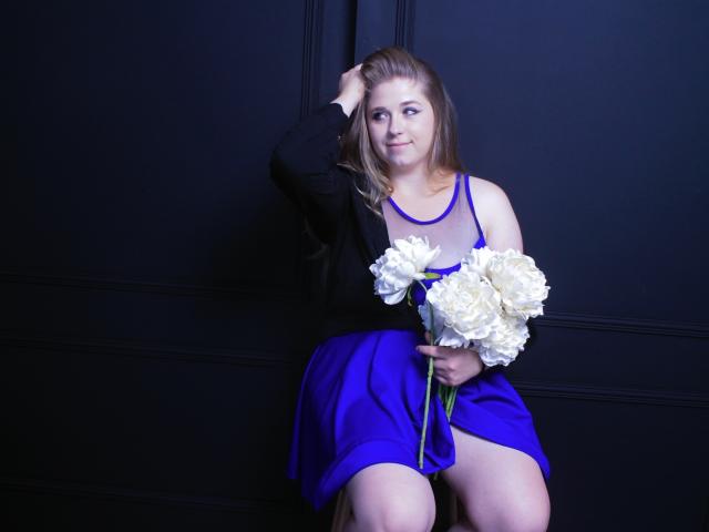 JessikaDaniels with blonde hair, black top, and blue dress, holding white roses, sitting against black wall, right hand in hair, smiling, blue eyes, fair skin, medium tits.