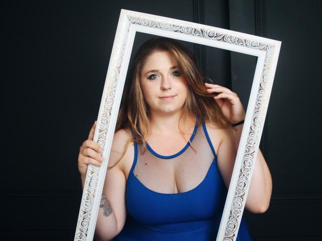 JessikaDaniels with medium tits, blue dress, holding ornate white frame, standing against dark background, light brown hair, subtle smile, right hand in hair, left hand on frame.