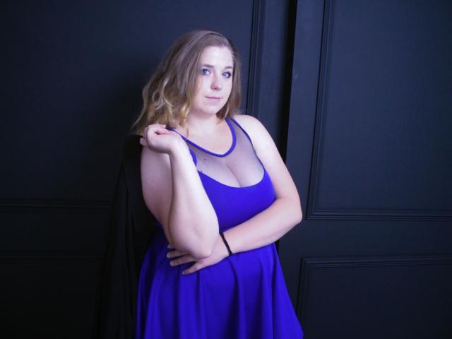 JessikaDaniels, de figura voluptuosa con grandes senos, vestida con un vestido azul, tiene el cabello castaño ondulado, está de pie frente a una pared negra, con una mano en la cadera y la otra tocándose el cabello, sonriendo suavemente..