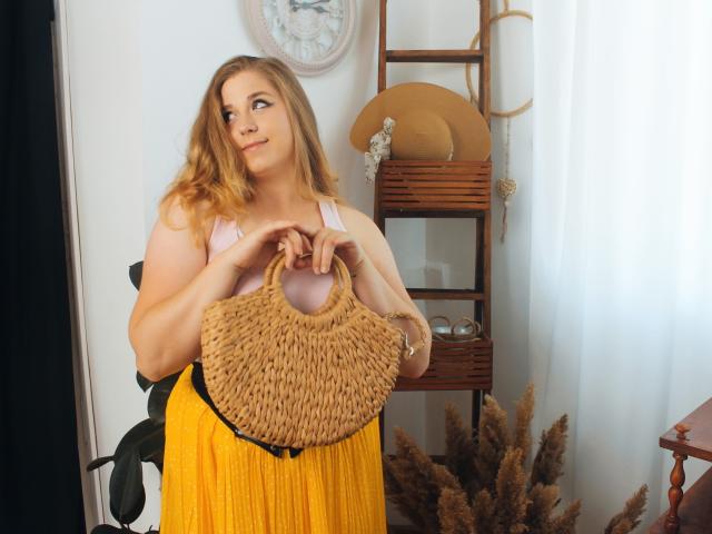 JessikaDaniels with wavy blonde hair, pink tank top, and yellow skirt, holding a woven tan bag, looking to the side, standing in a sunlit room.