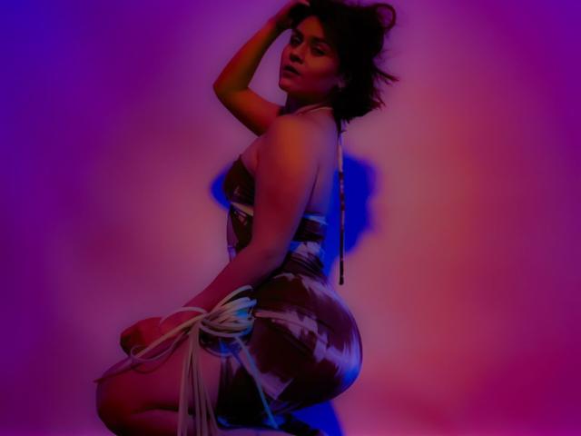 IsabelaCooper with dark hair, wearing a tight, patterned dress, and white rope on her ass, kneeling, hand in hair, illuminated by blue and pink lights.