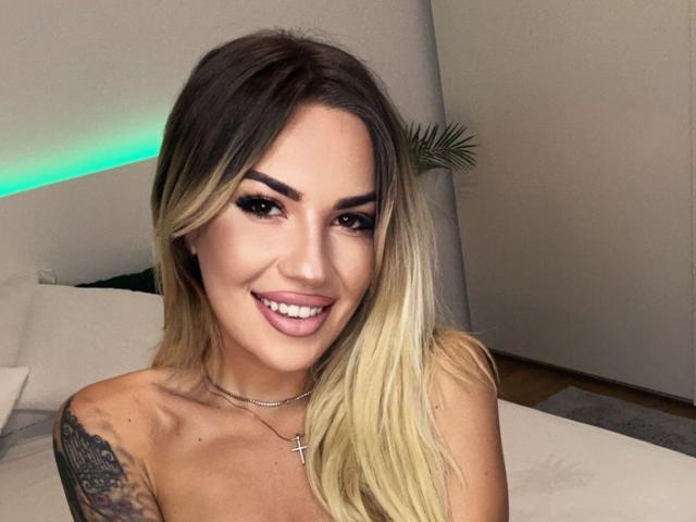 ZoeyMarie has blonde, ombre hair, dark eyebrows, a cross necklace, and a tattoo on her right shoulder. She's smiling with pink lipstick, showing her teeth.