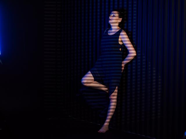 ElizabethSo stands barefoot, wearing a black dress, with one leg raised and hands on her hips, lit by striped shadows, against a dark, ribbed wall.
