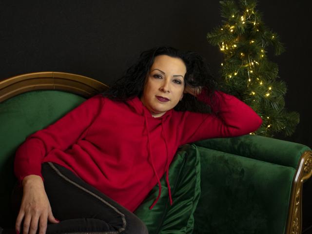 ElizabethSo with curly black hair, wearing a red hoodie and black pants, lounging on a green velvet couch, her hand behind her head, Christmas tree with lights to the right.