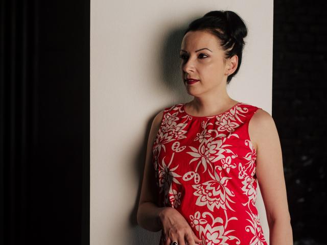 ElizabethSo with dark hair in an updo, wearing a red floral dress, standing against a white wall, looking down with a slight smile, red lipstick, holding her tits.