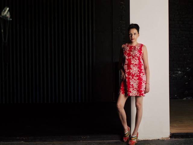 ElizabethSo stands against a white column, wearing a red floral dress, green and red heels, and a serious expression, with her ass slightly raised and her legs crossed.