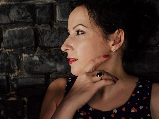 ElizabethSo with red lipstick, black hair, wearing a dark dress with heart patterns, touching her chin with a ringed hand, showing off her red nails and ear studs.