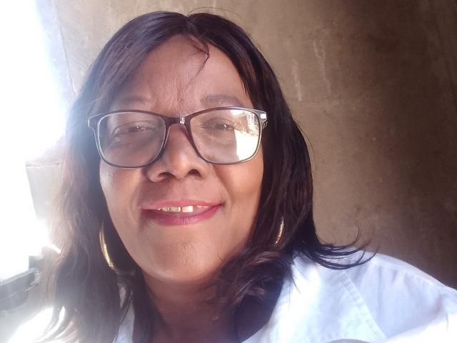 SexyBoobsNassX with black hair, glasses, white shirt, gold earrings, and pink lipstick, smiling with teeth showing, sunlight on left side.