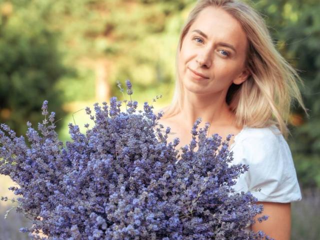 VasilisaSexy with blonde hair, holding a big bush of purple lavender, wearing a white shirt, smiling slightly, in a sunny, green, blurry background.