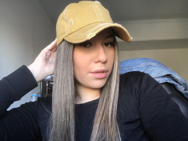 AlexaLip with long straight brown hair, wearing a black long-sleeve shirt and tan baseball cap, touching her hat with her right hand, looking at the camera.