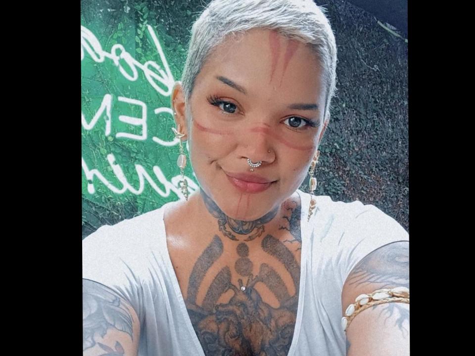 CarollyneD with short, grayish-blonde hair, nose ring, and tattoos, smiling, blood on face, white shirt, gold bracelet, and tattoos on chest and arm.
