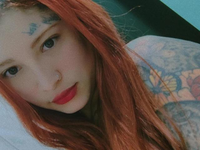 NinaNinnaS has red hair, tattoos on her shoulder, blue eyes, red lips, and a nose ring. She's looking at the camera with a slight smile.