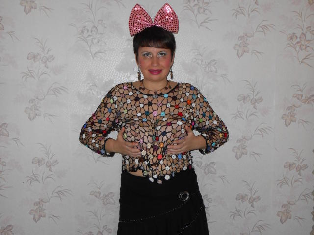AirMagdalene met kort bruin haar, die een rode strik met pailletten draagt, met haar kleurrijke top met mozaïekpatroon, een zwarte rok en een ketting, staand voor een bloemen behang..