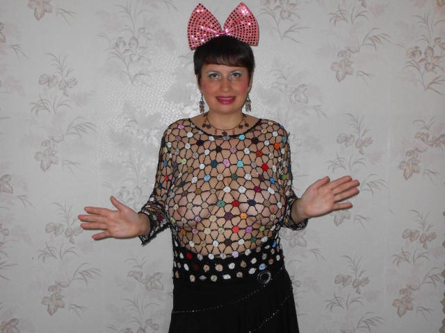 AirMagdalene con grandes senos, vistiendo una blusa transparente con estampado, un lazo de lunares rojos y blancos, una falda negra y un cinturón de cadena, sonriendo con los brazos extendidos, de pie frente a....