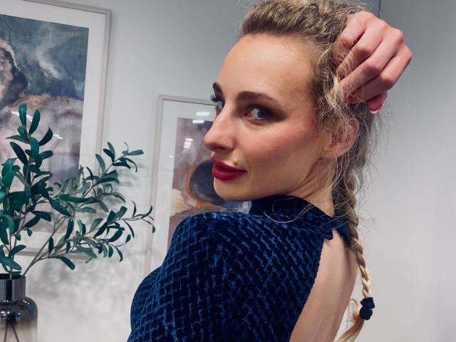 MarilynPretty with blonde braid, red lipstick, blue patterned dress, looking over shoulder, hand in hair, subtle smile, green plant on left, framed art on white wall.