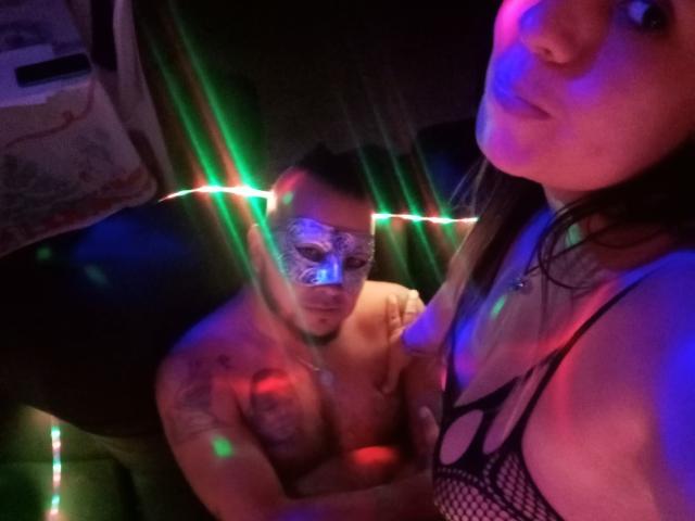 GoodsOfTemptation with masked, tattooed dude and his bare chest, her fishnet top, colorful lights, and her pouty face, in a dimly lit, sexy, intimate setting.