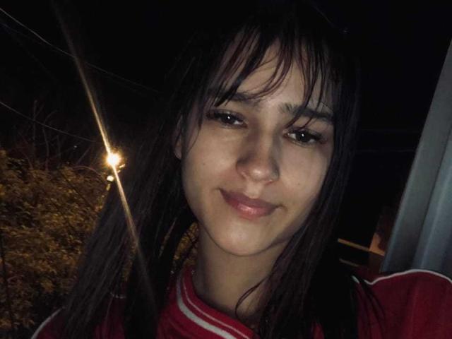 MelodyMansson has dark brown hair, a cute nose, and is smiling with pink lips. She's wearing a red and white shirt, looking cute in the dim light.