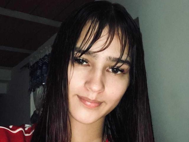 MelodyMansson has dark, straight hair with bangs, light brown skin, and a subtle smile. She has thick eyelashes, small nose, and is wearing a red and white top.